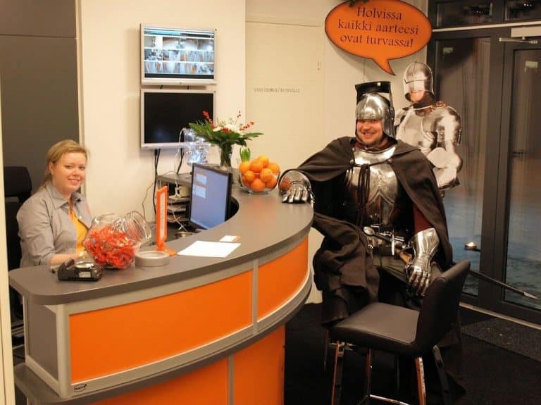 Holvin toimiston oranssi palvelutiski. Tiskin vieressä seisoo ritarihaarniska.