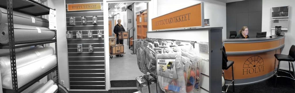 Mustavalkoinen kuva Holvin myymälästä: seinällä riippulukkoja ja muuttotarvikkeita, taustalla asiakas työntää kärryllä laatikoita.